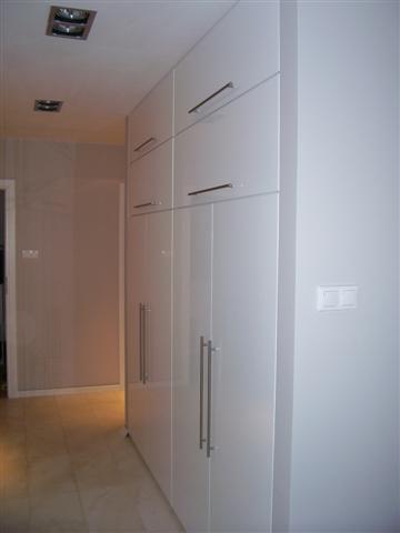 Biała, wysoka szafa z połyskiem i długimi, srebrnymi uchwytami, zamontowana na korytarzu z beżowymi ścianami i podłogą z płytek.